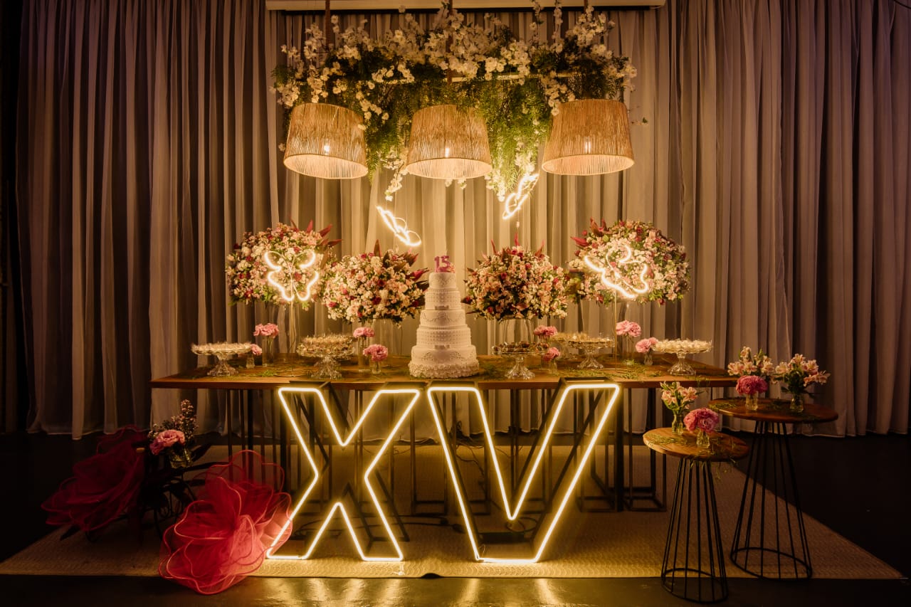 Mesa do bolo com letreiro XV em neon, arranjos florais e borboletas iluminadas