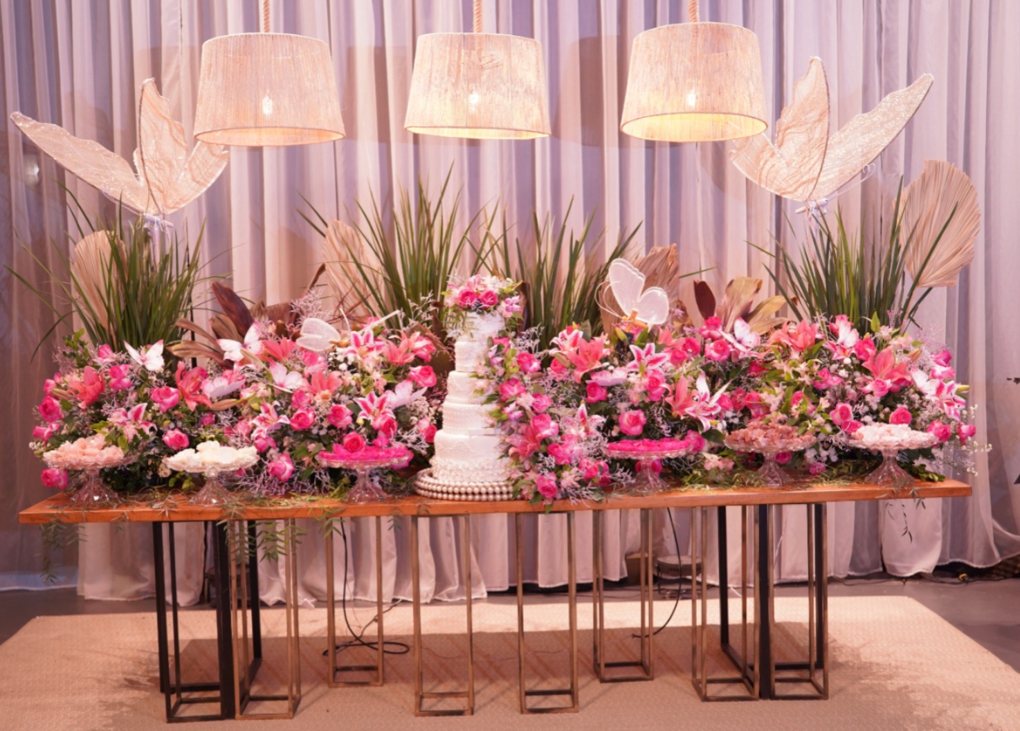 Mesa do bolo de 15 anos decorada com lírios e rosas pink, borboletas e luminárias de fibra no Jardins Caiçara